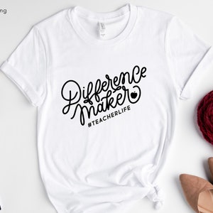 Op de afbeelding: Wit T-shirt met zwarte tekst die "Difference Maker" zegt met een rood appelpictogram en de hashtag "#TEACHERLIFE" eronder.