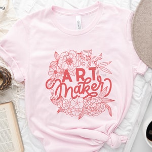May include: Pink t-shirt with a floral design and the text "Art Maker" in red.
