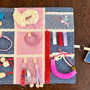 Puede incluir: Un tablero de actividades sensoriales con varias texturas y elementos. Presenta una tela azul estampada con cuadrados rosas, botones, cremalleras, cintas, una pulsera, una goma para el pelo, llaves y una pequeña almohada azul con la palabra "NAME".