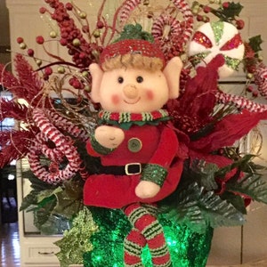 May include: A Christmas elf figurine wearing a red and green outfit sits on top of a green glittery hat with a black base. The elf is holding a candy cane in its hand.