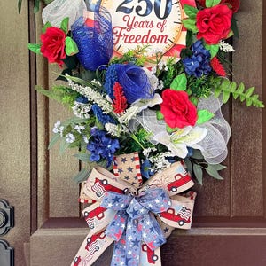 May include: A patriotic wreath with red, white, and blue flowers, greenery, and a large bow. The wreath features a circular sign that reads "250 Years of Freedom". The bow has a star pattern and red truck design.
