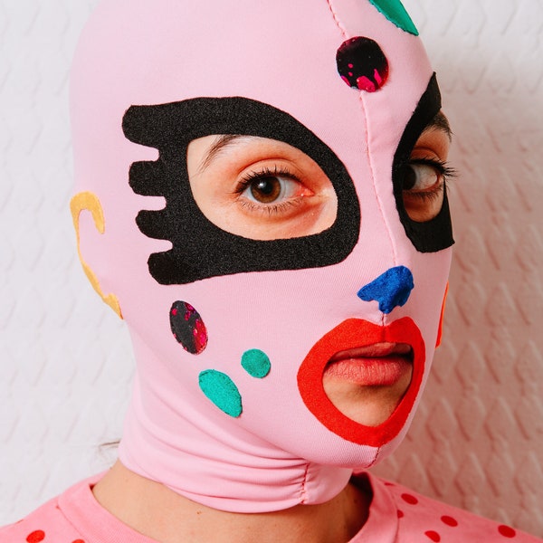 Soft pink mask with colorful dots, red lips, yellow right ear shape and big black eye contours.