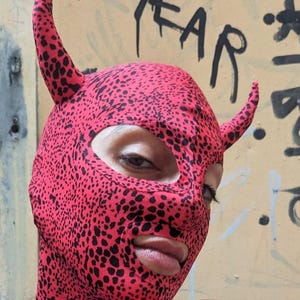 May include: A red and black spotted balaclava with horns. The balaclava covers the head and neck, with openings for the eyes and mouth. The word "TEAR" is visible in black graffiti on the wall behind the balaclava.