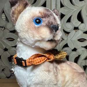 Può includere: Un peluche a forma di gatto siamese con pelliccia color crema, orecchie, muso e zampe marrone scuro e occhi in plastica blu brillante. Il gatto indossa un collare in tessuto a quadri arancione e marrone con un piccolo papillon e un campanello.