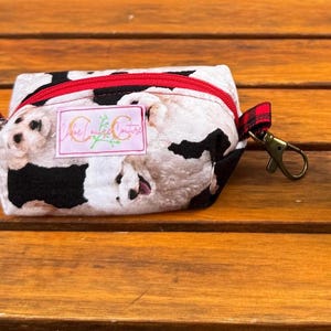 Peut inclure: Une petite pochette rectangulaire zippée avec un motif répétitif de chiens blancs et noirs. La pochette possède une fermeture éclair rouge, une étiquette blanche avec le texte Chloe Couture et un mousqueton en métal sur une patte à carreaux.