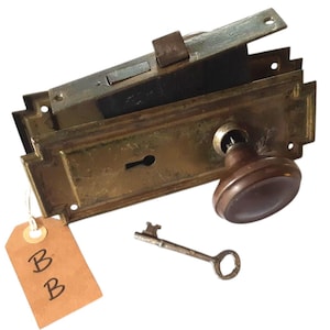 May include: A brass mortise lock set featuring a rectangular faceplate, a dark brown round knob, and a keyhole. A metal skeleton key lies below the lock mechanism. A tan rectangular paper tag with the handwritten letter B is attached to the lock.