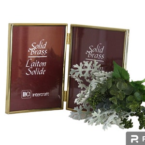 May include: A hinged double photo frame made of polished gold-tone metal with a thin border. The burgundy inserts feature the white text Solid Brass, Laiton Solide, and the IIC Intercraft logo. A small bouquet of green and silver foliage sits in front.