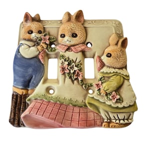 May include: Ceramic light switch cover featuring three bunnies, one in blue overalls, one in a pink dress, and one in a green dress. The bunnies are holding flowers and the cover has a floral design.
