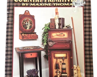Può includere: Un libro intitolato "COUNTRY PRIMITIVES 9" di Maxine Thomas. La copertina presenta un collage di mobili e decorazioni in stile country primitivo, tra cui un armadio, un tavolo e una mensola a muro, il tutto in toni caldi e rustici. Il libro è rilegato con un motivo a quadri.