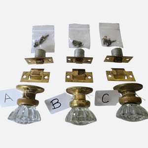 May include: Three sets of clear glass door knobs with brass bases, each labeled with a letter A, B, or C. Each set includes a metal latch mechanism, a brass strike plate, and a small plastic bag containing mounting screws for installation.