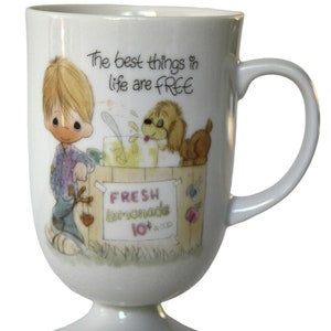 Peut inclure: Mug blanc en céramique avec un pied et une anse. La tasse présente une illustration de dessin animé d'un garçon et d'un chien vendant de la limonade. Le texte sur la tasse se lit « Les meilleures choses de la vie sont GRATUITES » et « LIMONADE FRAÎCHE 10¢ LE VERRE ».