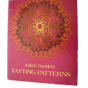 May include: A softcover book titled Tatting Patterns by Julia E. Sanders with a magenta cover featuring a circular orange and brown tatting design. The top left corner displays the Dover Needlework Series logo and a small white price sticker.