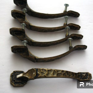 May include: Five antique brass cabinet door pulls with a hammered finish and a decorative design. The pulls have a curved shape and are attached to a flat back plate with screws.