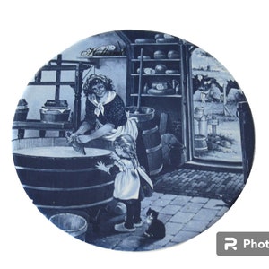 May include: A blue and white decorative plate featuring a pastoral scene. The image depicts a woman and child in a dairy setting, with cows visible in the background. The word "Holland" is written at the top of the plate.