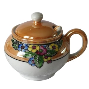 May include: A small, brown ceramic sugar bowl with a lid and a handle. The bowl is decorated with a floral pattern in shades of blue, yellow, green, and pink.
