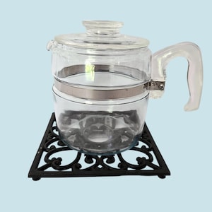 May include: A clear glass coffee pot with a lid and handle, held together by a metal band. The pot sits on a black, ornate, square trivet. The handle is clear and curved.