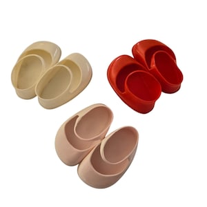 May include: Three pairs of doll shoes in different colours: cream, pink and red. The shoes are made of plastic and have a simple design.