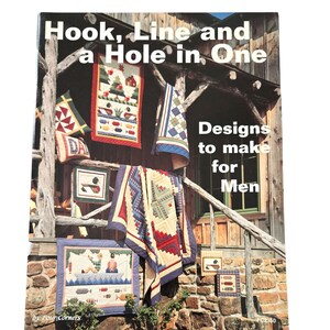 Op de afbeelding: Een boek getiteld "Hook, Line and a Hole in One: Designs to make for Men" door Four Corners. De omslag toont een rustieke houten veranda met een verscheidenheid aan kleurrijke quilts en wandkleden met vis- en buitenmotieven.