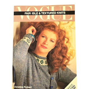 May include: A book titled "Vogue Fair Isle & Textured Knits" by Christina Probert. The cover features a woman wearing a blue knitted sweater with a white and green Fair Isle pattern. The book is part of the Vogue Knitting Library.
