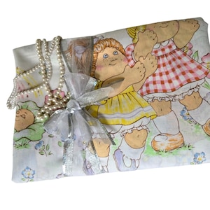 May include: A white fabric gift bag with a silver bow and a pearl necklace. The bag has a print of three dolls with flowers.