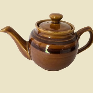 May include: A brown ceramic teapot with a lid and handle. The teapot has a curved spout and a rounded body. The color is a mix of light and dark brown, with a glossy finish. The teapot is designed for brewing tea.