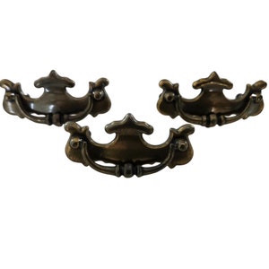 May include: Three antique brass cabinet pulls with ornate designs. Each pull has a curved shape with a decorative center and scrollwork on the sides.