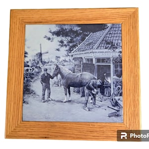 May include: A framed blue and white ceramic tile depicting a scene of a blacksmith working on a horse's hoof in a rural setting. The tile is surrounded by a wooden frame.