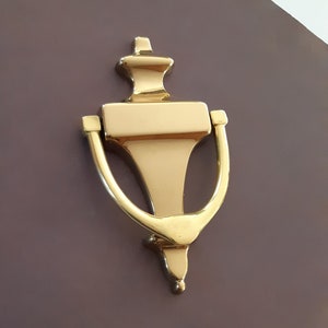 May include: A gold door knocker with a decorative design. The knocker is shaped like a bell with a rounded top and a curved handle.