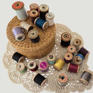 May include: A selection of vintage wooden thread spools in diverse colours, such as white, black, blue, and purple. The spools are arranged on a woven basket and a cream lace doily, creating a nostalgic craft theme.