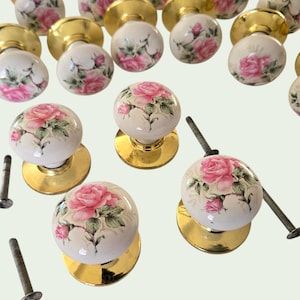 May include: A collection of vintage door knobs with a white porcelain finish, adorned with pink rose floral patterns. The knobs have gold-toned bases and are accompanied by screws, suggesting a hardware or home decor theme.