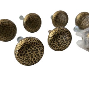 Peut inclure: Ensemble de sept boutons de meuble de couleur bronze avec un motif texturé et martelé. Les boutons sont ronds avec une petite vis transparente. Un ensemble est encore dans son emballage plastique d'origine. Idéal pour la rénovation.