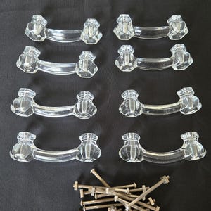 Peut inclure: Huit boutons de cabinet en verre transparent avec un design facetté et une finition argentée. Les boutons sont disposés en quatre rangées de deux. Un tas de vis argentées se trouve au centre inférieur de l'image.