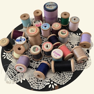 May include: A selection of vintage thread spools in a variety of colours, including blue, purple, green, and pink. The spools are made of wood and cardboard, and are arranged on a black, lacy doily. The spools vary in size and shape, some with labels.