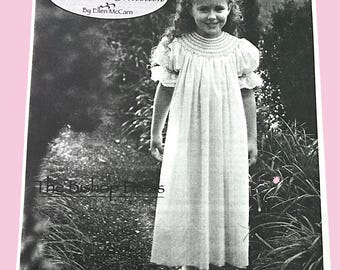 May include: Black and white image of a young girl wearing a long, white dress with a smocked bodice and short, puffed sleeves. The dress is part of "The Classic Heirloom Collection" by Ellen McCann. The girl is standing in a garden setting.