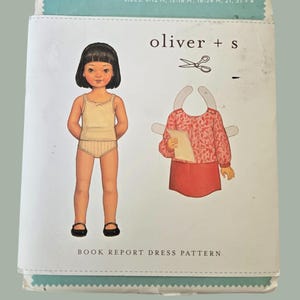 Peut inclure: Un patron de couture avec une illustration de poupée en papier. Le paquet porte le texte "oliver + s" et "BOOK REPORT DRESS PATTERN". L'illustration montre une poupée en sous-vêtements et un modèle de robe. Les tailles sont indiquées comme 6-12 M, 12-18 M, 18-24 M, 2T, 3T + 4.