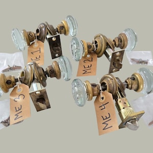 May include: Four vintage glass door knobs with brass fittings. Each knob features a clear, oval glass handle and is connected to a brass base. Each knob has a tag with the letters "ME" and a number. Small bags of screws are also included.