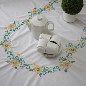May include: White tablecloth with a floral cross-stitch design in yellow and green. A white teapot, four teacups, and a small green plant in a white pot are arranged on the cloth, creating a cozy tea setting.