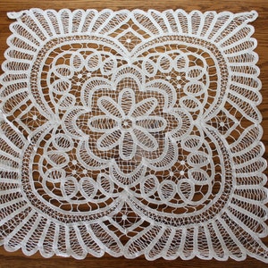 May include: A square, white lace doily with an intricate floral and geometric design. The doily is placed on a wooden surface, showcasing its delicate openwork and detailed patterns. The lace features a central flower motif and scalloped edges.