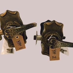 May include: Two antique bronze door handles with ornate detailing. Each handle has a decorative backplate, a curved lever, and a keyhole. The handles are labelled "Ypp 1" and "Ypp 2" on small tags. Hardware and screws are included.