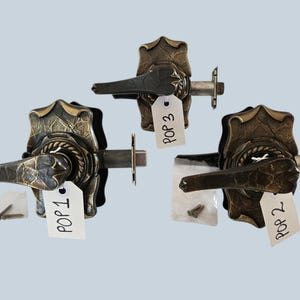 May include: Three antique bronze door handles with intricate leaf and rope detailing. Each handle has a lever and a decorative backplate. Small bags of screws and tags labeled "POP 1", "POP 2", and "POP 3" are included.