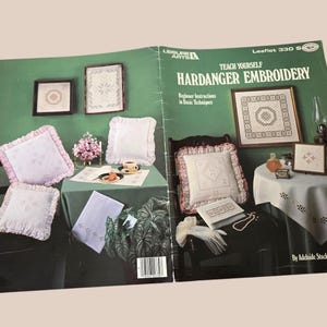 Peut inclure: Un livre vintage intitulé "Teach Yourself Hardanger Embroidery" avec des instructions pour débutants. La couverture présente des échantillons de broderie encadrés et des coussins brodés dans un cadre domestique. Le livre est ouvert, affichant la couverture et les pages intérieures.