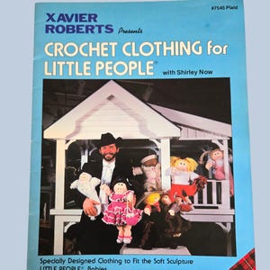 May include: A book cover with the title "CROCHET CLOTHING for LITTLE PEOPLE" by Xavier Roberts. The cover features a man holding dolls wearing crochet clothing. The background is a light blue with a plaid design in the corner.
