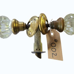 May include: A vintage door knob set featuring two clear, fluted glass knobs with brass bases and circular brass rosettes. A central brass latch mechanism connects the knobs. A brown paper tag with the handwritten text #002 is attached.