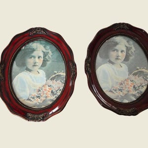 May include: Two oval antique picture frames in a dark red colour. Each frame contains a vintage portrait of a young girl in a white dress, holding flowers. The frames feature ornate, decorative detailing.