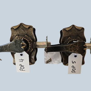 May include: Two antique bronze door handles with ornate floral designs. Each handle has a lever and a decorative backplate. The handles are labeled "POP 4" and "POP 5" on white tags. Small screws are included.