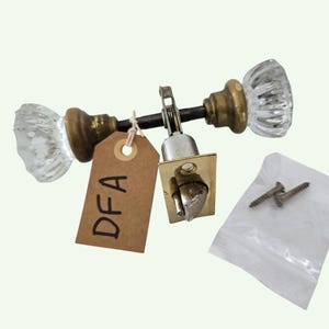 May include: Antique door hardware set featuring two clear glass doorknobs with brass bases, a brass-colored latch, and a small bag of screws. A brown tag with "DFA" is attached to the hardware.