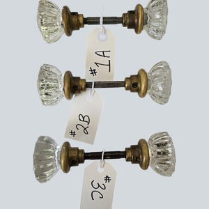 May include: Three glass door knobs with fluted, clear glass handles and antique brass-toned metal bases. Each knob is connected by a central metal spindle and features a white paper tag labelled #1A, #2B, and #3C respectively, attached by string.