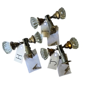 May include: Four antique glass door knobs with brass hardware. Each knob has a clear, faceted glass design and is attached to a metal plate. Small white tags with the letters "H" and "J" are attached to some knobs. Screws are included.