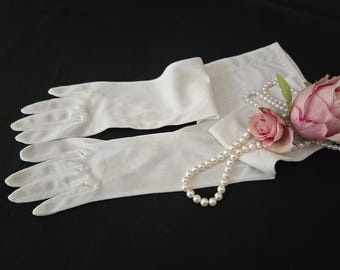 May include: A pair of elegant, off-white gloves with delicate stitching, laid out on a black surface. A pearl necklace and two pink roses with green leaves are arranged near the gloves, creating a vintage aesthetic.