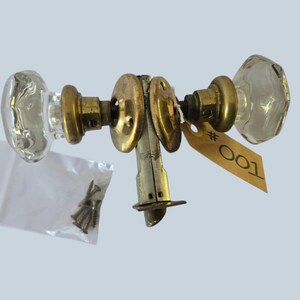 May include: A vintage door knob set featuring two clear, faceted glass knobs attached to brass hardware components. The assembly includes a central latch mechanism, a small plastic bag containing four metal screws, and a tan paper tag labelled #001.